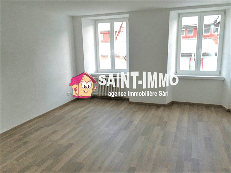 Empty room with white walls, wooden floor, and two windows. A radiator is mounted on the wall below the windows.