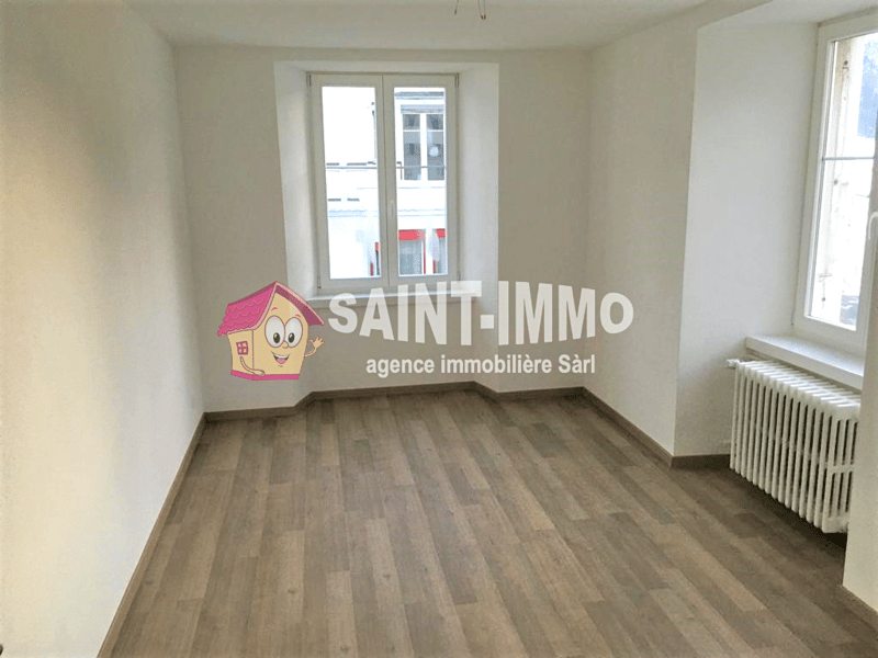 Empty room with white walls, wooden floor, and two windows. A radiator is installed on the right wall.