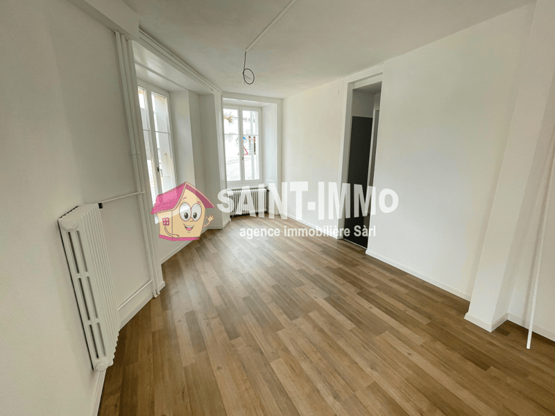 Empty room with white walls, wooden floors, and white radiators. Two windows with white frames and white doors. A watermark is present in the image.