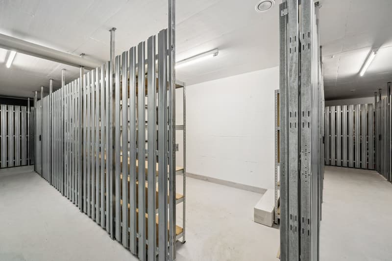 The image shows a large storage room or warehouse with rows of metal shelving units or racks. The room has a white, minimalist design with bright lighting. The shelving units are made of metal and appear to be used for storing various items or materials.