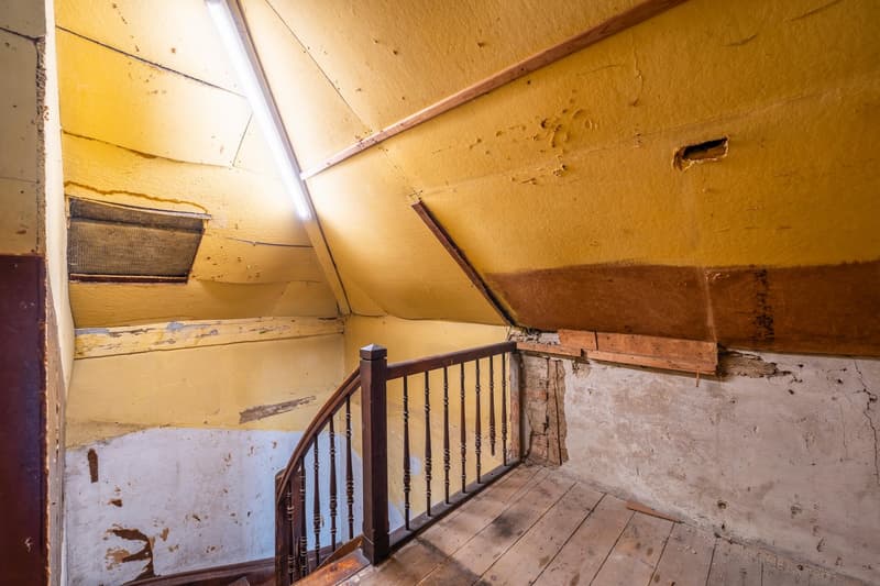 Interior of house showing attic space with wooden railing, worn yellow and white walls, wooden ceiling with visible beams, and damaged parts.