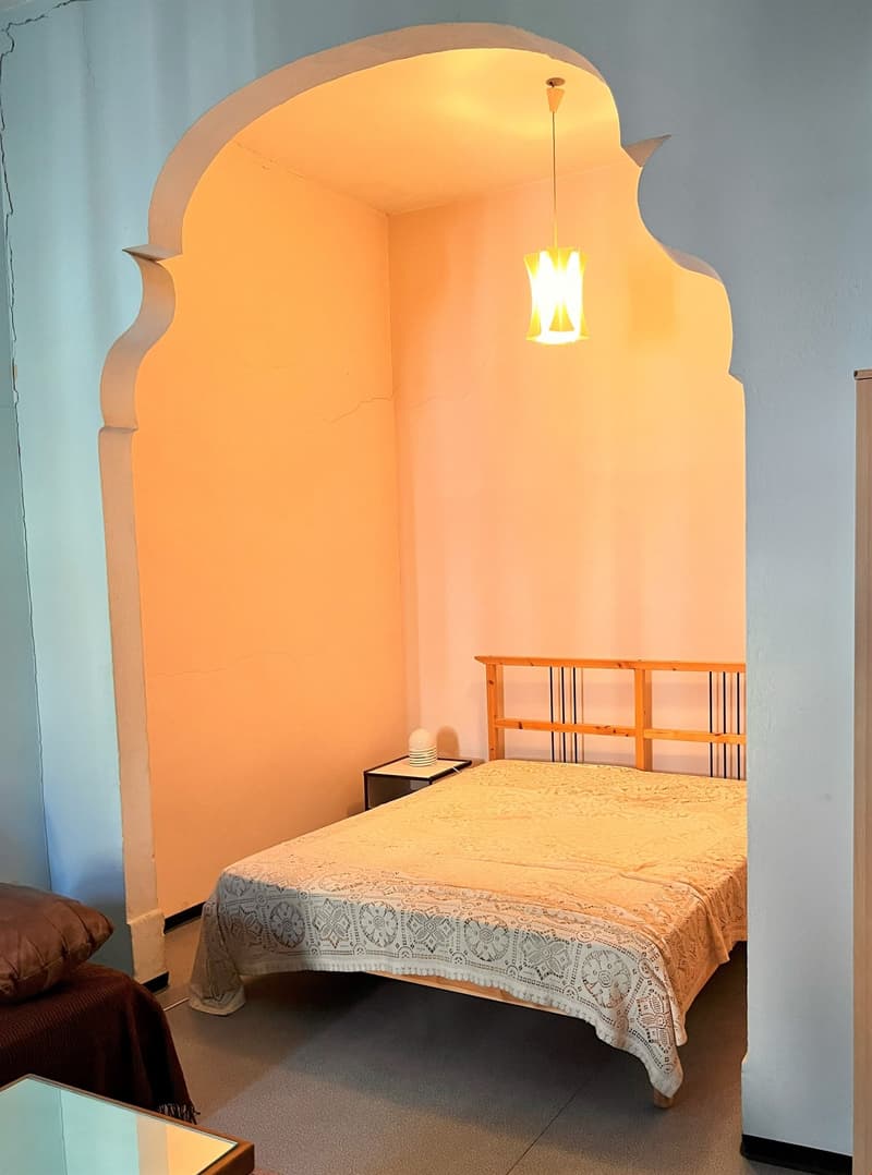A bedroom with a wooden bed headboard, bedspread, bed pillow, night lamp, nightstand, carpet, and an archway in the corner.