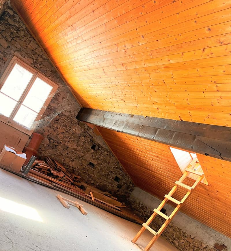 A wooden ceiling, a wooden ladder, a wooden window with glass, a stone wall, wooden pieces, and a small cardboard box on the floor