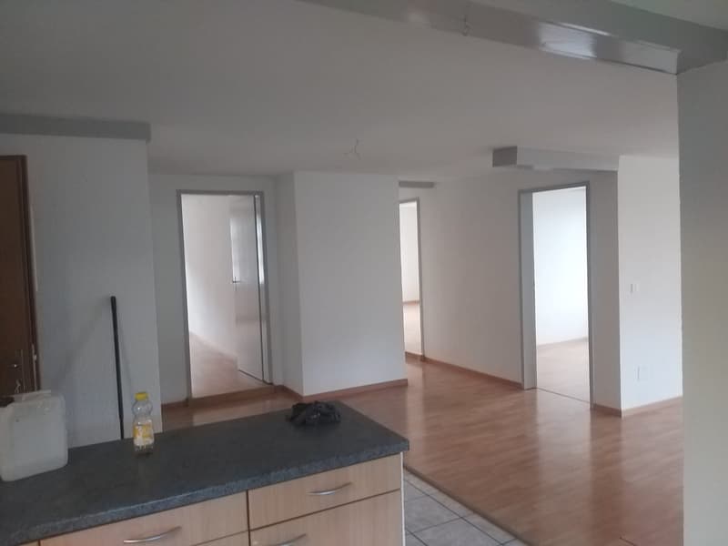 Empty room with wooden floor, white walls, kitchen counter, cabinets, and doors leading to other rooms