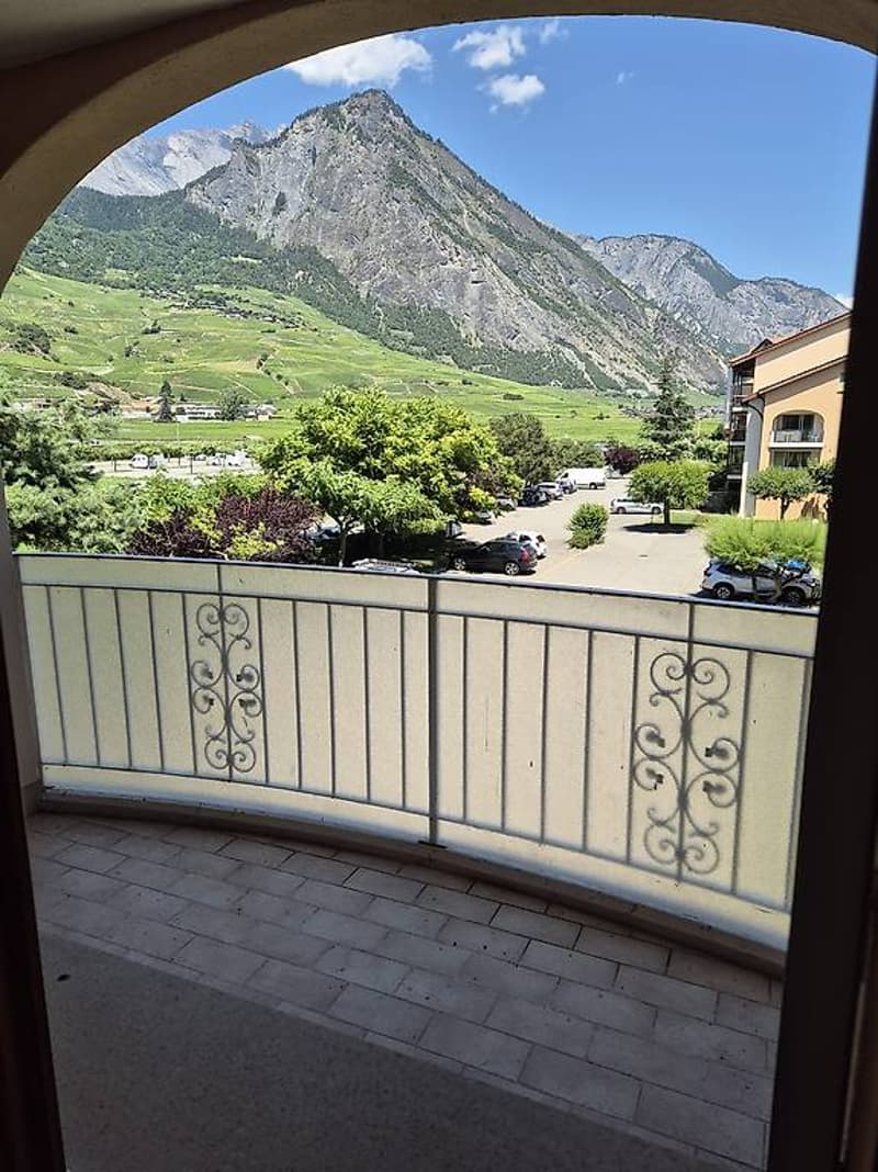 Balcony with mountain view, tiled flooring, railing, trees, parked cars, distant houses, greenery