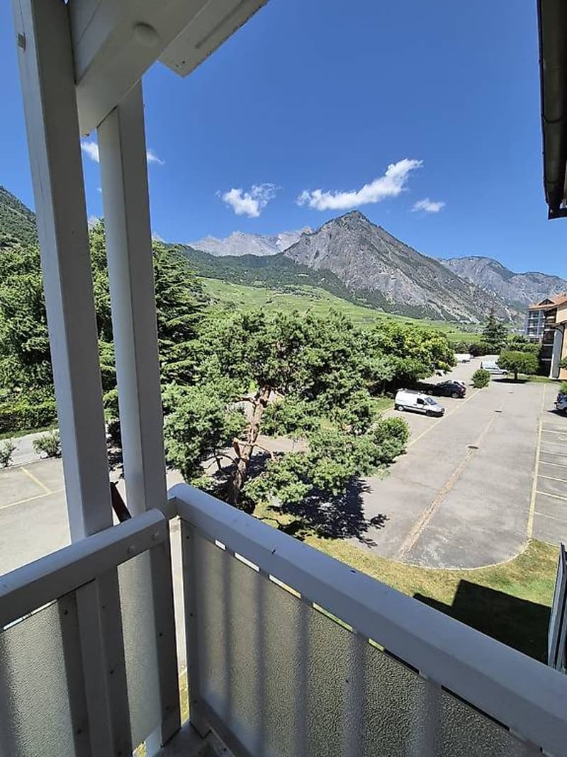 White railing balcony with transparent panel, car parking lot with cars, lush trees, building, mountains