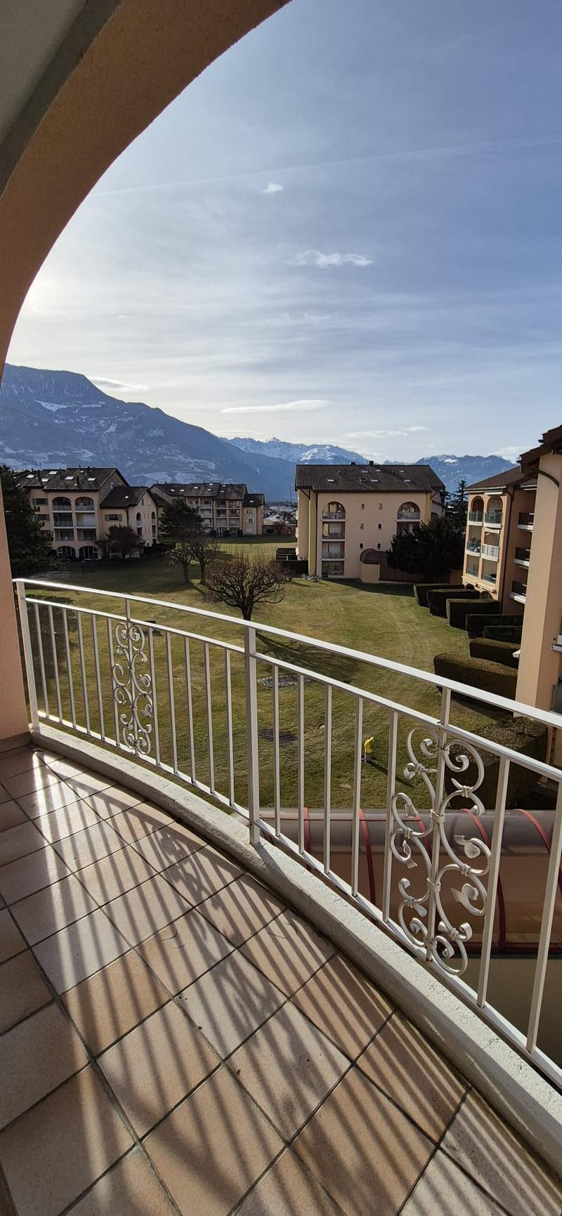 A balcony with a view of a grass field, a small hill, mountains in the background and some apartment buildings.