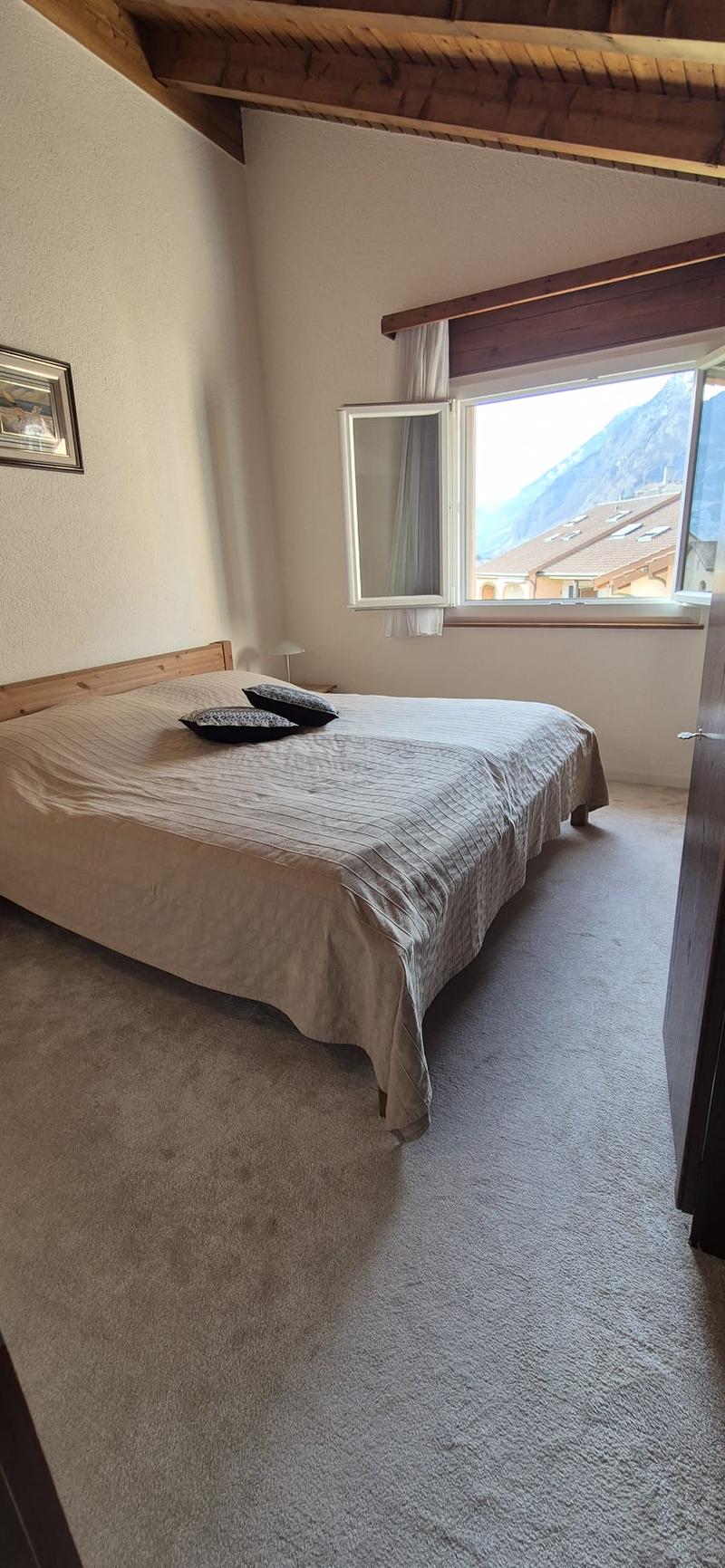 A room with a large double bed in the middle, two pillows, a lamp on the side table, white curtains, wooden ceiling, and a view of the mountains outside