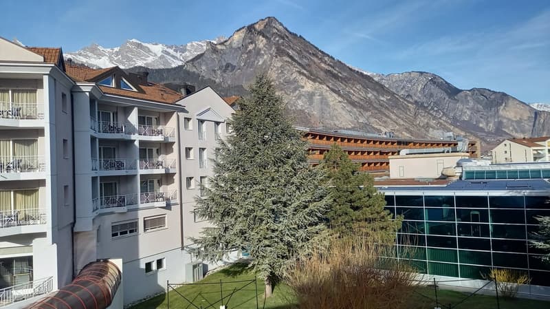 A multi-story building with balconies, surrounded by trees and grass, with a mountain in the background.