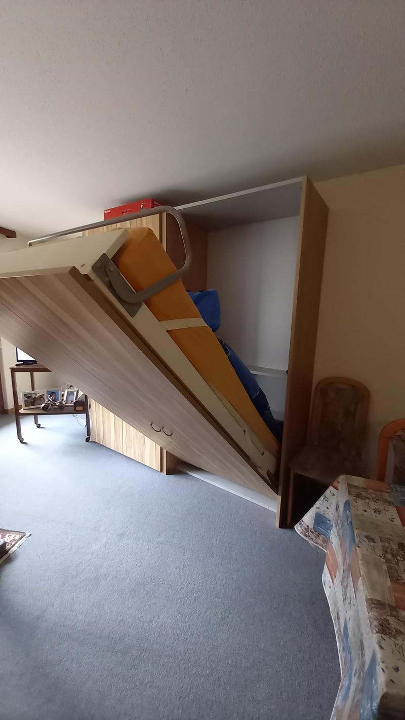A room with a pull-out bed, a wooden cabinet, a table with picture frames, and a chair.