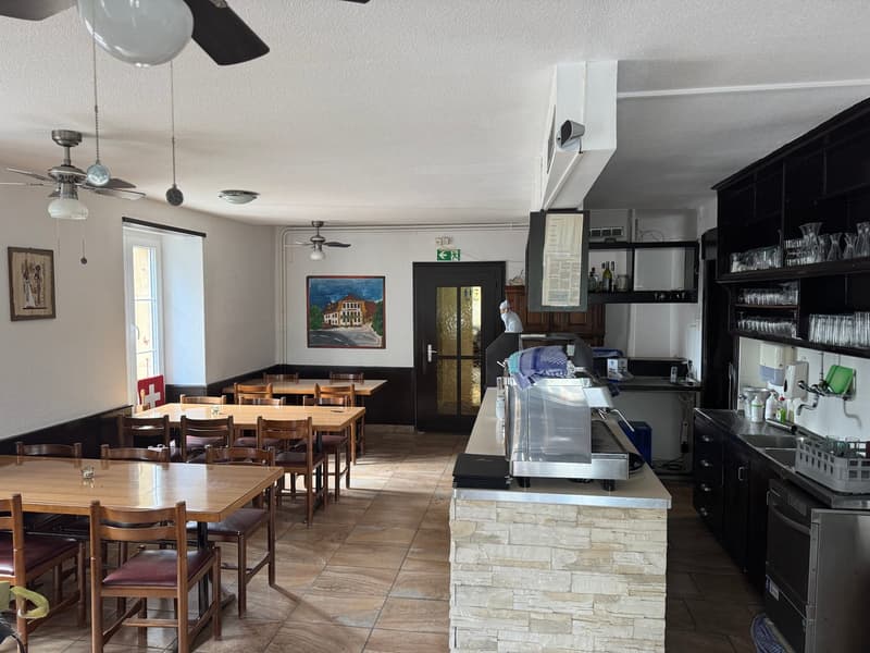 The image shows the interior of a restaurant or cafe. There are several wooden tables and chairs set up for dining. The kitchen area is visible in the back, with a stone counter and various kitchen appliances and equipment. A dishwasher is visible on the