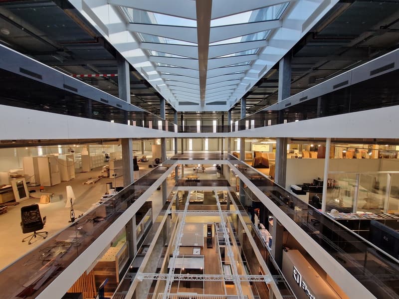 Multi-level atrium, steel and glass framing, offices and desks in multiple floors, white walls, natural light from skylights, multiple floors with offices, desks, chairs, and other office supplies.