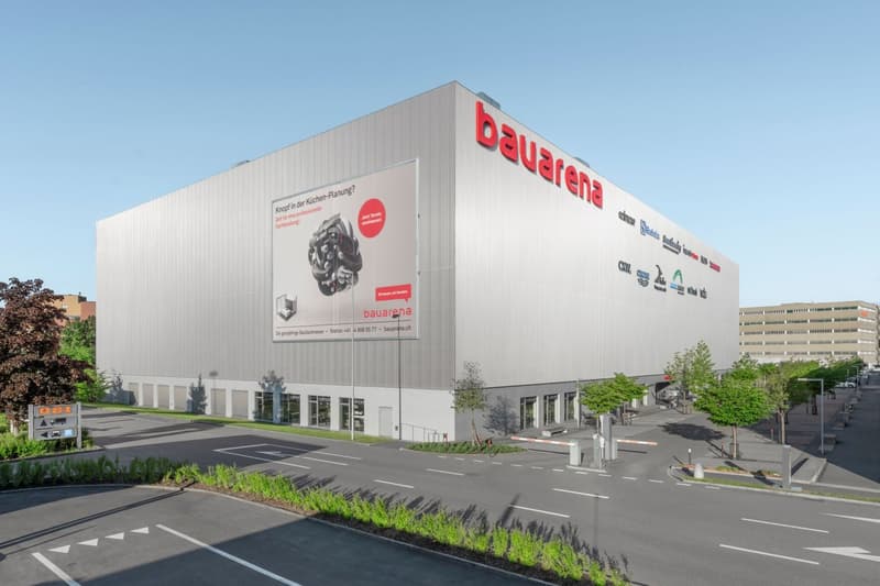 Large industrial building, gray exterior, large advertisement banner, multiple entrance doors, extensive parking area, city backdrop