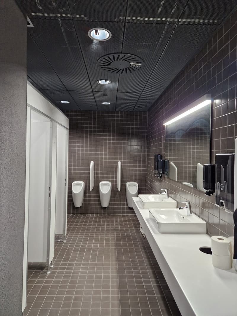 Public restroom with urinals, sinks, and mirrors. Tiled flooring, white walls, and ceiling-mounted fixtures.