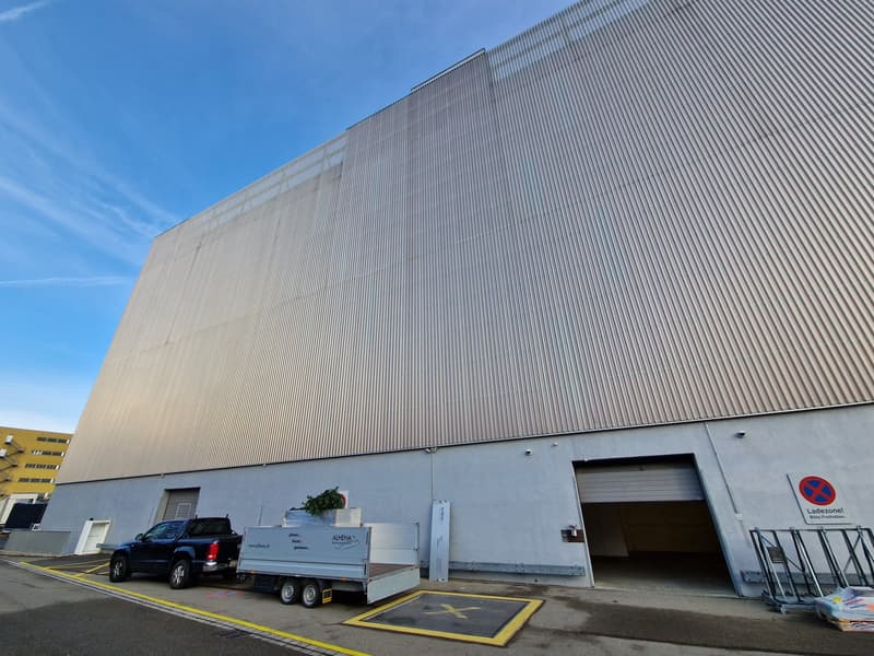 Large metal facade, grey wall panels, large open garage, black pickup truck with trailer parked in front, yellow line markings, metal handrails on side, signboard near garage door