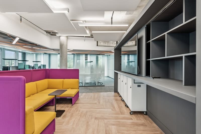Wooden floor, glass walls, vibrant colored sofas, table, storage shelves, white cabinets, large windows