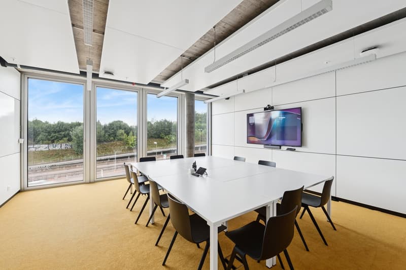 white table, black chairs, large glass windows, mounted TV, white walls, brown flooring