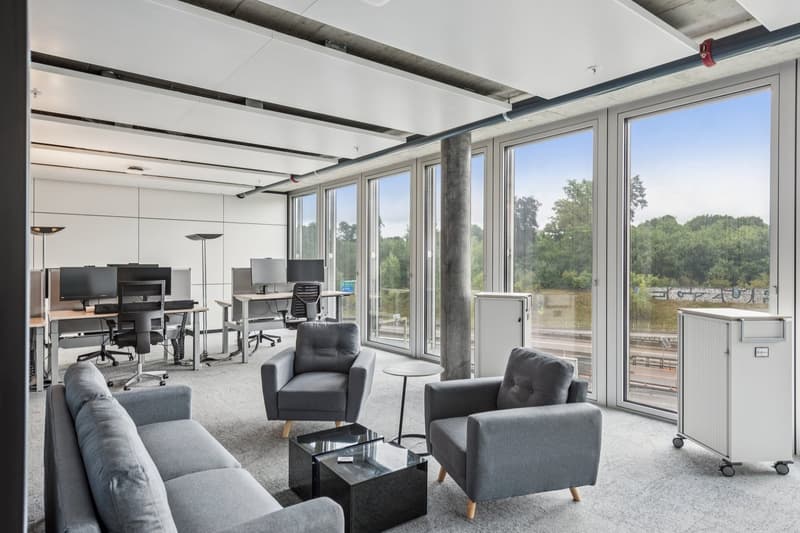 Modern open office space with gray chairs, sofas, desks, and large windows. Desks have monitors and office supplies. Central coffee table, small side table, and a movable cabinet.