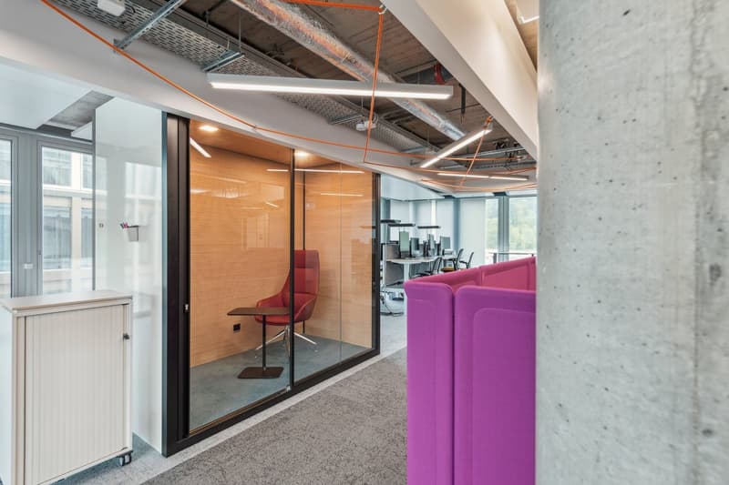 Modern meeting room with glass walls, office furniture, grey carpet, ceiling lights, and red and purple cubicles.
