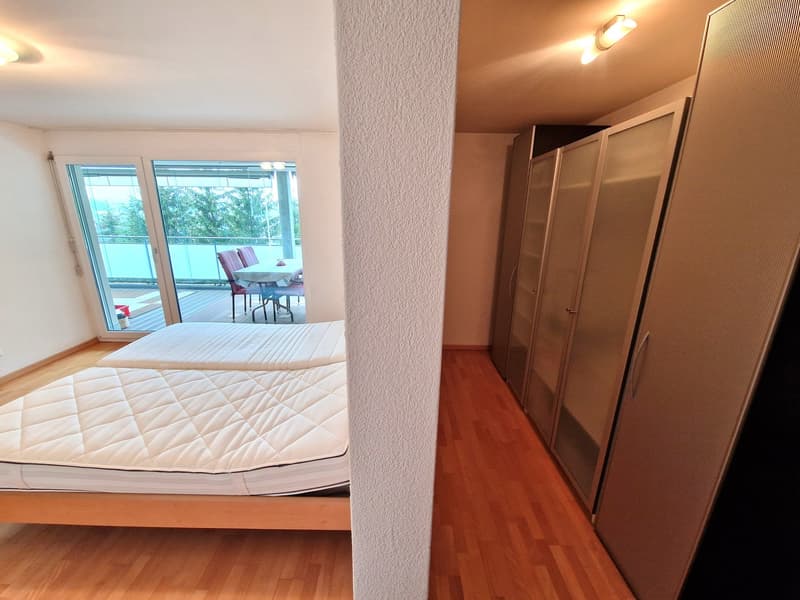 Spacious bedroom with large windows, hardwood flooring, and a balcony. The room features a queen-sized bed with a quilted comforter and a wardrobe cabinet.