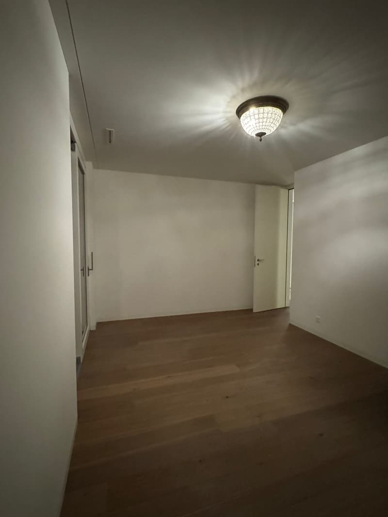 An empty room with a hardwood floor, white walls, a ceiling light, and an open door.