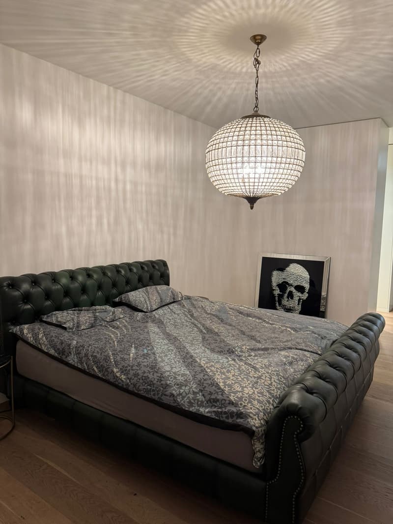 A bedroom with wooden floor, large bed, bed covers, pillows, bedside table with metal frame, wall-mounted framed artwork with skull print, chandelier on the ceiling