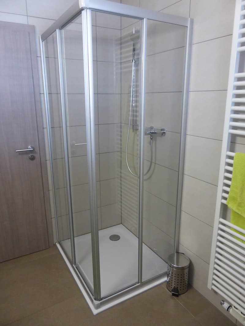 A modern bathroom with a glass shower cubicle with a chrome-plated shower head, white tiled walls, and tiled floor. A small trash can is located near the wall, and a towel rack is attached to the wall.