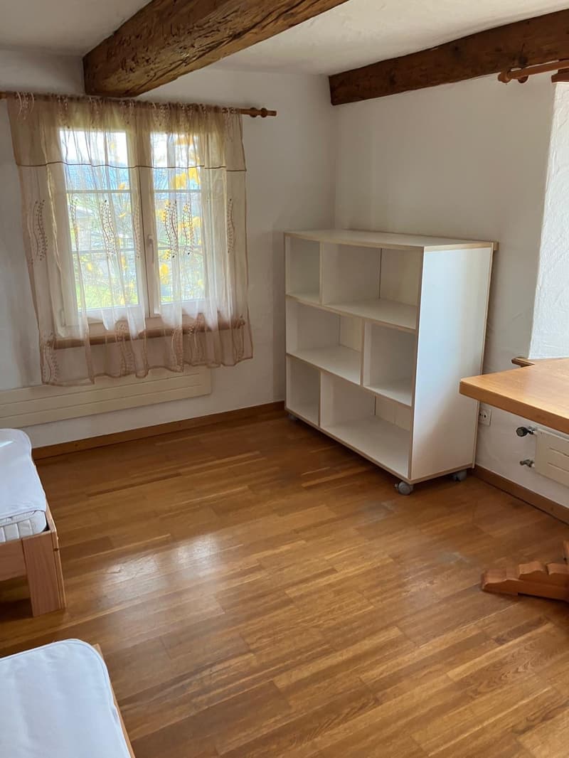 small bedroom with wooden floors, white walls, single bed, window with curtains, shelf, and wooden desk