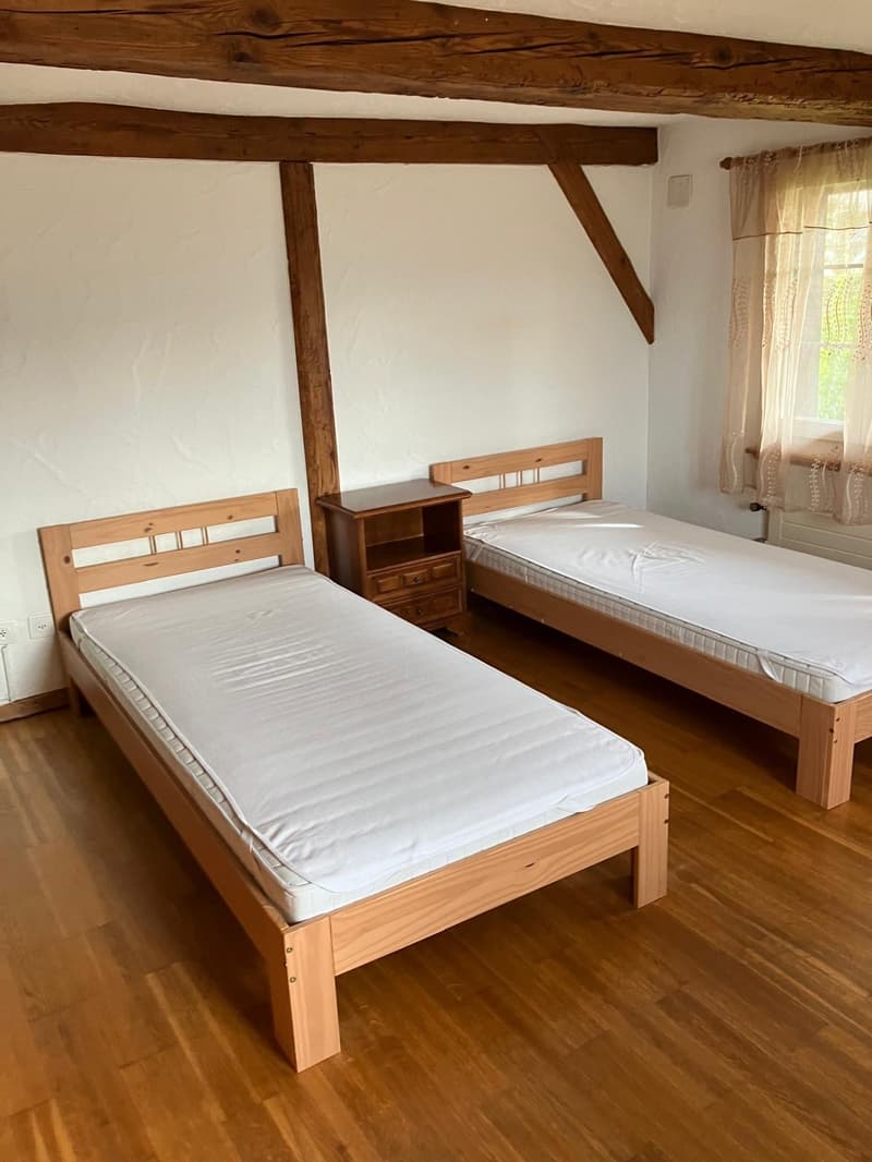 a bedroom with two beds and a side table, wooden flooring, white walls, wooden beams, curtain on window