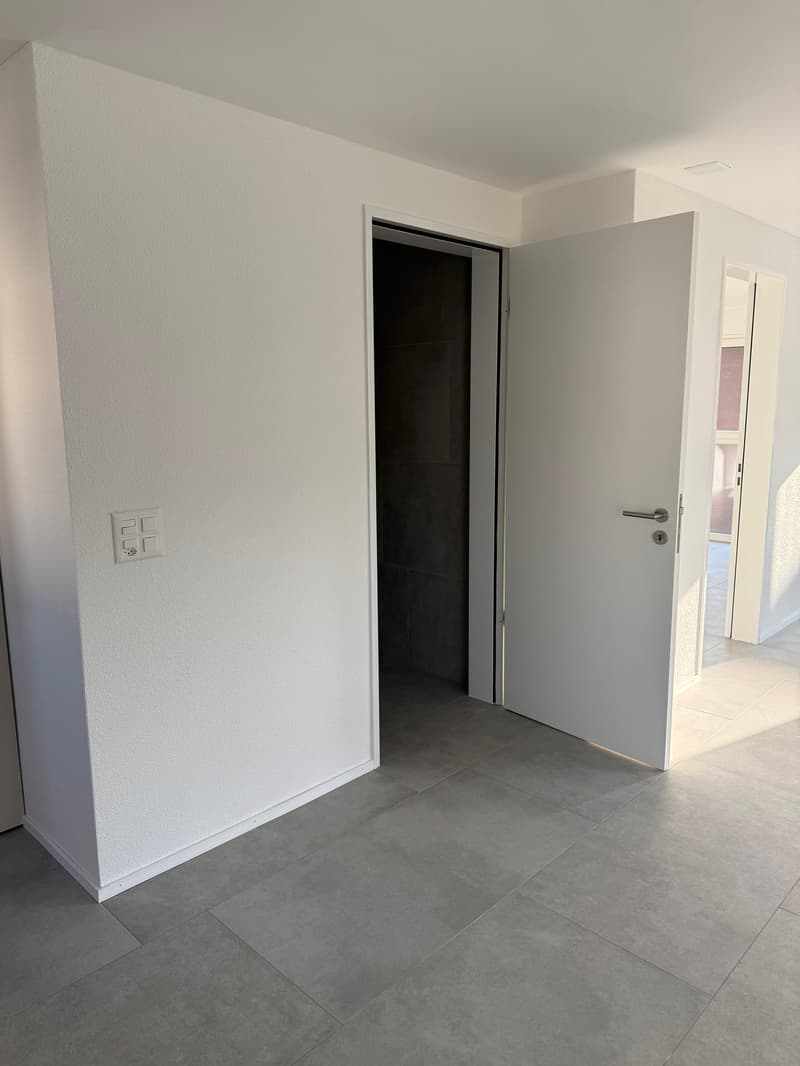 Empty room with white walls, gray floor, open door, power outlet on the wall