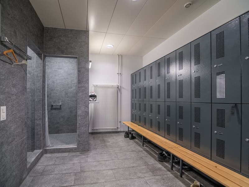 Locker room with gray walls, tiled floor, wooden bench, multiple lockers, a shower stall, and a radiator.