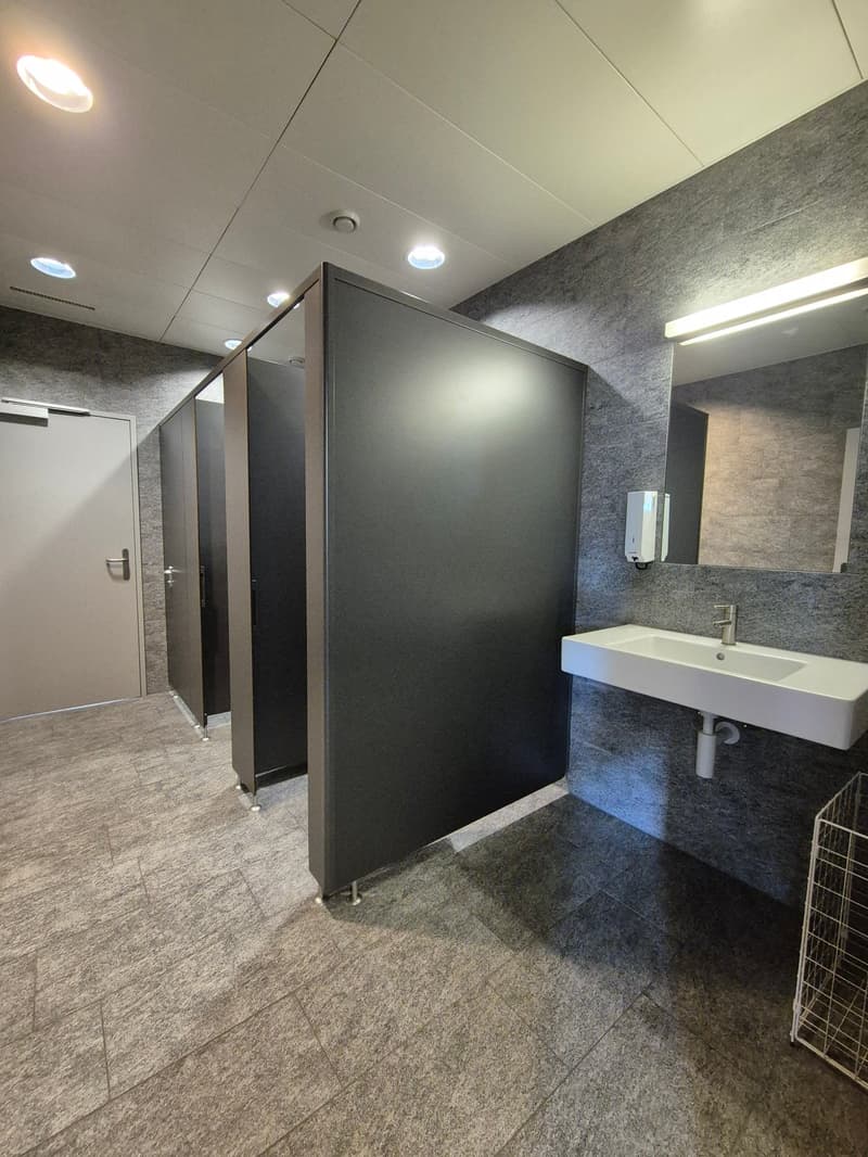 3 toilet stalls, grey walls, recessed lighting, large mirror, white sink, soap dispenser, baskets