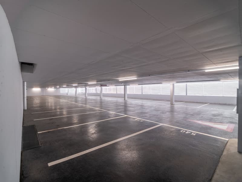 Empty underground parking lot with white painted parking spaces, concrete floor, fluorescent lighting, and large windows.