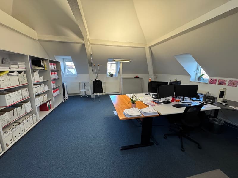 A modern office with wooden desk, two chairs, three monitors, computer keyboard, book shelves, blue carpet, windows.