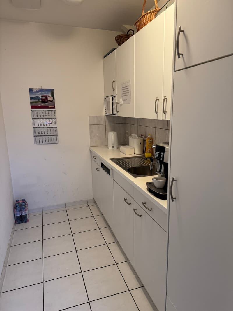 Modern kitchen with white cabinets, tiled floor, stainless steel sink, black countertops, microwave, kettle, toaster, and cleaning supplies.