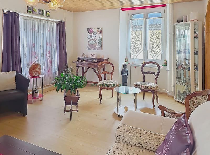Living room with wooden floor, white walls, large windows, wooden desk, chairs, glass table, potted plants, decorative items, and a couch with pillows.