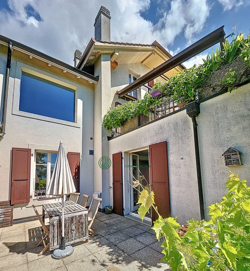 Cozy patio area with wooden table and chairs, plants and umbrella, brick wall, brown wooden doors, balcony with railings and plants