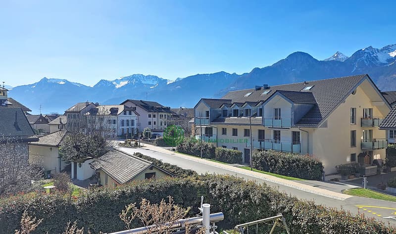 Aerial view of a residential area with several houses, mountains in the background, and a road with greenery on the sides.