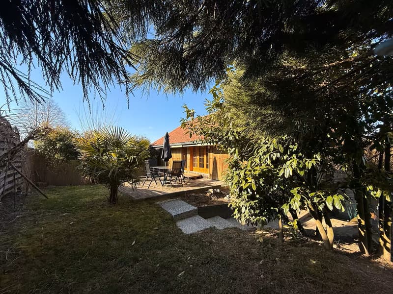small detached wooden house, red roof, wooden front deck, outdoor dining table and chairs, closed umbrella, gravel pathways, enclosed by trees and plants