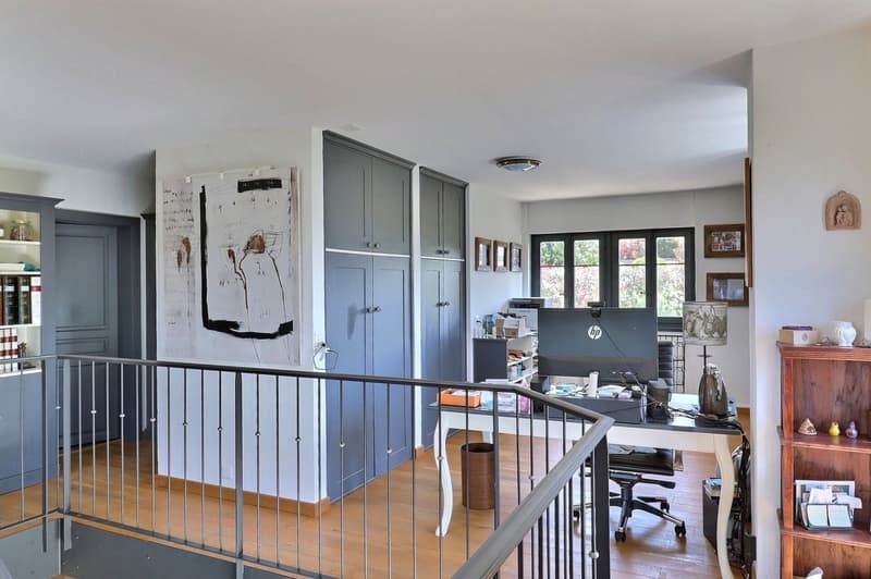 Wooden floor, desk with computer, office chair, lamp, bookshelf, cabinets, picture frames, railings, and wall decorations