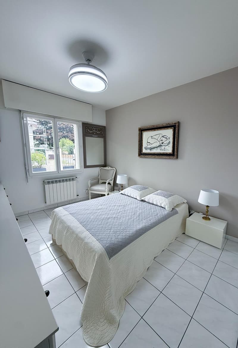 Modern bedroom with white tiles, gray walls, bed, lamp, mirror, picture frame, chair, and radiator