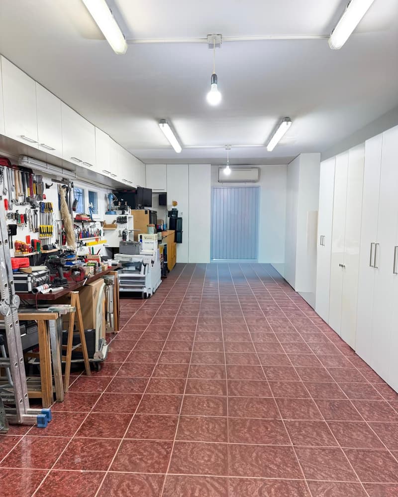 Garage with red tiled floor, white cabinets, tools on shelves, ceiling lights, and equipment.