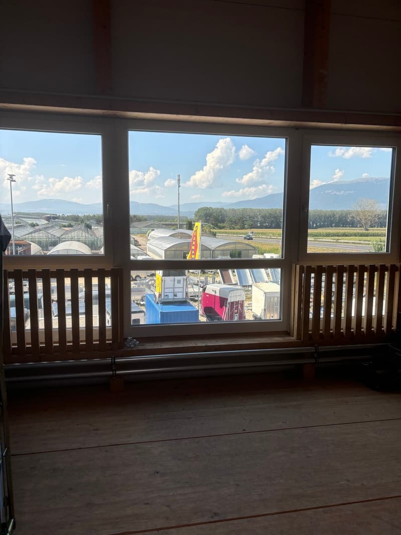 The image shows a room with large windows overlooking a scenic outdoor landscape. The windows provide a panoramic view of mountains, forests, and a commercial or industrial area with various structures and vehicles visible in the distance.