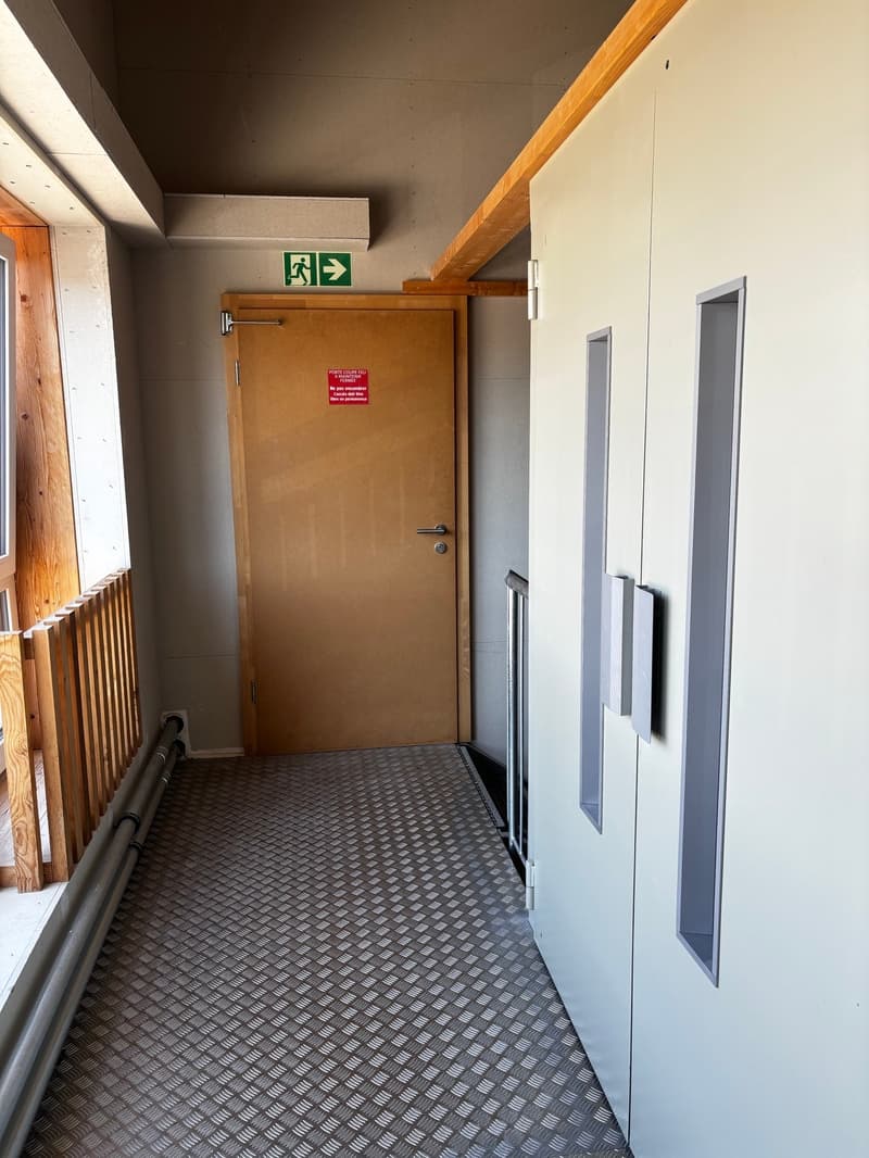 Empty hallway with wooden door, metal floor, white walls, and a window