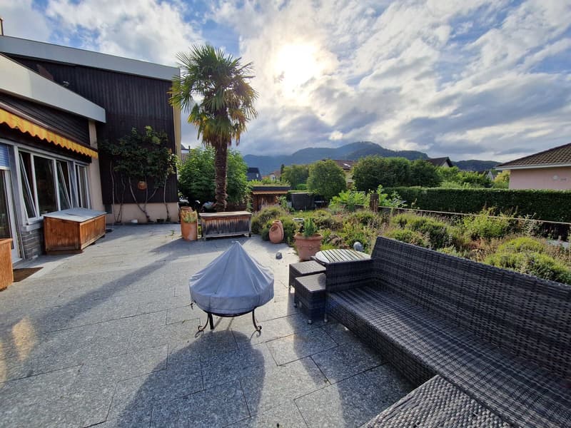 The image shows an outdoor living area with a patio, wicker furniture, a fire pit, and a palm tree. The space is surrounded by lush greenery and has a view of mountains in the background.