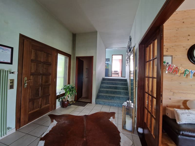 The image shows the entryway of a home. It has a wooden front door, a tiled floor, and a staircase leading up to the second floor. There is a plant and a decorative lantern in the entryway, as well as a 'Happy' sign on the wall. The walls are painted a li
