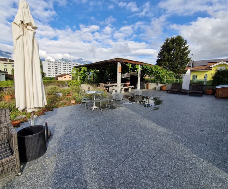 The image shows an outdoor patio area with a covered seating area, tables and chairs, and a large umbrella. The patio has a tiled floor and is surrounded by greenery, including a tree. There are buildings visible in the background, suggesting this is part