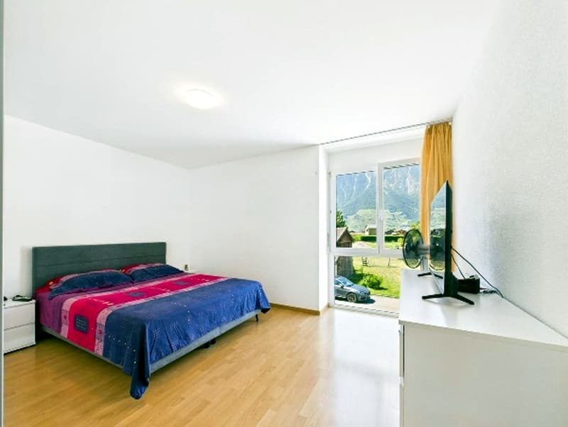 spacious bedroom, wooden floor, white walls, bed with bedding, bedside table, sliding glass door with view