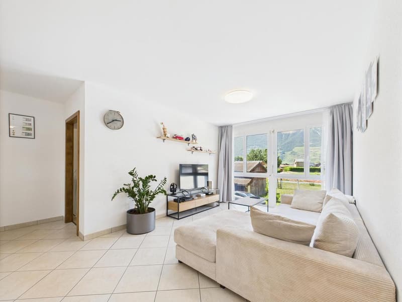 living room, white walls, flat screen TV, L-shaped sofa, potted plant, coffee table, floor lamp, large windows
