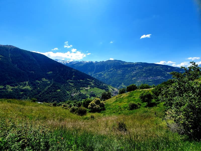 Mountains, lush greenery, clear blue sky with some clouds, distant town or village, wide expanse of land